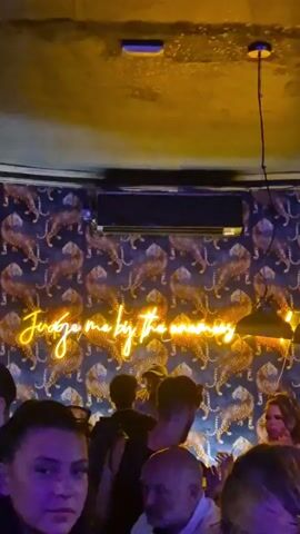 The video shows the inside of a busy bar. The walls are covered in a wallpaper featuring images of tigers. A neon sign reads "Judge me by the money." People are gathered around a bar, where a bartender is preparing drinks. The lighting is dim, with pendant lights hanging from the ceiling. Loud music is playing.