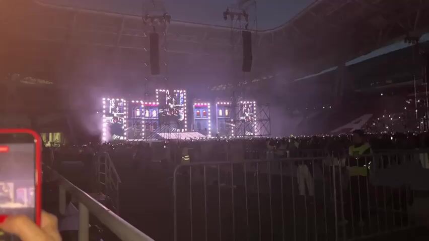 The video shows a live concert from the perspective of someone in the audience. The stage is large with multiple screens displaying visuals. There are many people in the audience, and some security personnel are visible. The lighting is mostly purple and pink, with smoke effects adding to the atmosphere. The sky is dark, indicating it is evening or night. A person's hand holding a phone is in the foreground, suggesting they are recording the event.