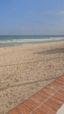 The video shows a static, medium-distance shot of a sandy beach on a sunny day. The sand is light brown and shows footprints. In the background, the ocean is visible, with gentle waves rolling towards the shore. The water transitions from a light turquoise near the shore to a deeper blue further out. The sky is mostly clear with a few wispy clouds.  A red brick walkway or border is partially visible in the foreground along the bottom of the frame. A white plastic bucket and a red can-like object are visible on the sand. Shadows are cast on the sand, likely from objects or structures just outside the frame.