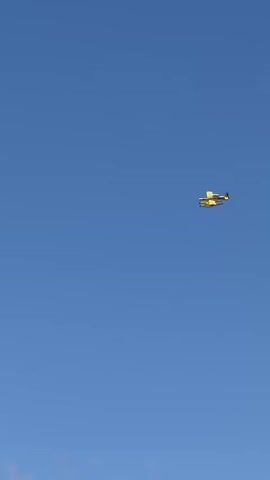 A yellow biplane flies across a clear blue sky. The plane is a small, single-engine aircraft with two sets of wings, one above the other. It appears to be a fixed-wing aircraft. The plane is seen from below and at a distance, making specific details about its markings or any occupants difficult to discern. The sky is mostly clear, with a few small, wispy clouds visible. The lighting suggests it is daytime, possibly mid-morning or afternoon. The plane is flying in a generally horizontal trajectory.