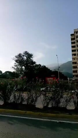 The video pans across a street scene with tall apartment buildings in the background. In the foreground, there is a median strip with bushes and yellow and black curb. A silver SUV drives past on the left, and other cars are visible on the road and parked in the background. The sky is bright and hazy.