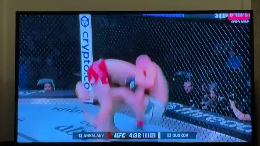 The media file shows a screen displaying a live broadcast of a mixed martial arts (MMA) fight. Two male fighters are grappling on the mat inside an octagon. One fighter, wearing red shorts, is on his knees, attempting to control the other fighter who is on top of him. The second fighter, with a bald head and wearing black gloves, is positioned behind the fighter in red, with his arms wrapped around his opponent. The cage surrounding the octagon is visible, as are spectators in the background. The lighting appears to be artificial, consistent with an indoor arena.

Visible text on the screen includes "UFC" along with a timer indicating "4:32" and fighter names: "ANKALAEV" and "GUSKOV." There is also text in Armenian at the top right of the screen, and "FAST SPORTS" logo. The scene is set in an MMA arena, and given the location context of Sevan, Armenia, it is likely this fight is being broadcast within that region. The activity depicted is a high-intensity grappling exchange during a professional MMA bout. The overall mood conveyed is one of intense physical struggle.