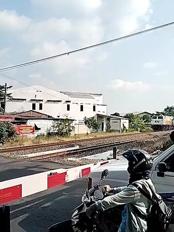 The video shows a daytime scene at a railway crossing in Sidoharjo, Indonesia, under a bright sky with scattered clouds. A silver and pink passenger train, pulled by a yellow and white locomotive marked "KAI" (Kereta Api Indonesia), is passing through the intersection. The railway crossing barriers are lowered, and the red warning lights are flashing, indicating the train's approach. Several vehicles, including a scooter with a person wearing a helmet and a car, are stopped, waiting for the train to clear the tracks. In the background, white buildings, some trees, and a large billboard with "Idul Fitri" visible text can be seen. The train moves from right to left across the frame.