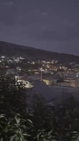 The video shows a cityscape at dusk. Lights twinkle in the buildings that spread across a hillside. The sky is a deep, dusky purple, with hints of clouds. In the foreground, dark green foliage obscures the bottom of the frame. A chimney with what appears to be a bird perched on top is visible in the middle ground.
