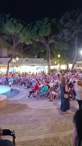 A large crowd of people are seated in an outdoor amphitheater at night, watching an event on a stage. The audience is composed of individuals of various ages, mostly adults, with many appearing to be elderly. They are seated in rows of folding chairs, facing a circular stage area that is partially visible on the left side of the frame. The stage appears to have some circular structures and is lit with spotlights. The audience members are casually dressed, and many are looking towards the stage. Behind the audience, there are trees with lush foliage, and in the background, there are buildings with illuminated windows and streetlights. The overall atmosphere suggests a public performance or gathering.