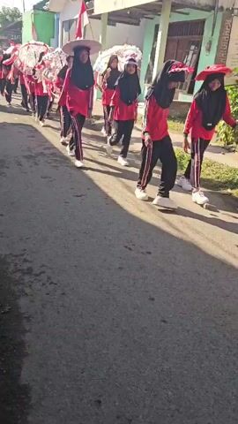 A group of young people, most of whom appear to be girls wearing hijabs and matching red shirts and black pants with red stripes, are participating in a parade or procession. They are walking down a paved street lined with buildings and houses. Some of the participants are holding large, decorative umbrellas adorned with red and white trim. Several Indonesian flags are visible, some attached to poles and others draped on buildings. The weather appears to be sunny. The group is moving from left to right in the frame. One individual in the foreground is wearing a wide-brimmed hat over their hijab.