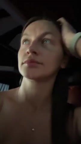 A woman is recording a video. She is in a dimly lit space, possibly inside a vehicle, as there is a dark ceiling visible behind her. She has medium-length brown hair, blue eyes, and is wearing a simple silver necklace. She is also wearing a white fitness tracker or watch. The woman's hand is resting on the side of her head as she looks at the camera and sings along to a song. The focus is primarily on her face.