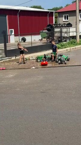 Four children are playing in a muddy area in front of a large red building and a house with a red roof. A young girl in a black top and skirt is standing on the left, looking down. A boy in a blue t-shirt and dark shorts is near the center, reaching out towards a boy in a red shirt who is lying on the ground. Two green toy tractors are also in the muddy area. Another green toy tractor is further to the right. The children appear to be interacting with each other and the toys. The environment is an outdoor setting with a paved road in the foreground and vegetation in the background.
