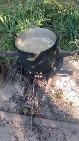 This is a first-person perspective video of someone cooking a large pot of food over an open fire. The pot is a dark, possibly cast iron, cauldron with two handles, and it's filled with a light-colored liquid and what appear to be pieces of food, possibly meat or vegetables. The pot is balanced on a makeshift stove constructed from metal bars and possibly old tires, situated over a pile of burning sticks and branches.

The scene is outdoors, with green foliage visible in the background, suggesting a rural or natural setting. The ground appears to be dirt or sand, with a scattering of twigs and ash from the fire. The lighting suggests it's daytime, with sunlight casting shadows.

The primary activity is the cooking process. The person recording is periodically interacting with the pot, using a ladle to stir or check the contents. In the first few frames, a ladle is seen entering the pot, followed by the person's hand reaching into the pot. The steam rising from the pot indicates the food is hot and actively cooking. The setting and method of cooking suggest a traditional or rustic approach.

There is no visible text in the video. The overall mood is one of simple, outdoor food preparation. The location context provided, Aragatsavan, Armenia, points to a specific region where such traditional cooking methods might be employed.