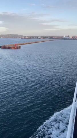 The video shows a wide shot of a body of water, likely the ocean, with gentle waves. In the foreground, the wake of a boat is visible on the right side of the frame. In the distance, a long, man-made structure, possibly a breakwater or pier, extends into the water. This structure appears to be made of interlocking segments and has red markers on it. Beyond the breakwater, a coastline is visible with buildings and structures, suggesting a populated area or a port. The sky above is partly cloudy with a soft, diffused light, indicating either dawn or dusk. The overall scene is tranquil and expansive.