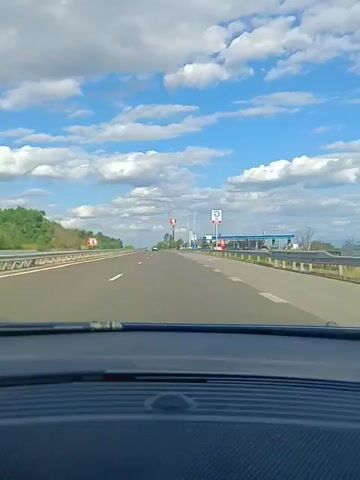 This is a first-person perspective video taken from inside a vehicle driving on a highway. The road is paved and has white lane markings. On the left side of the highway, there is a guardrail and then trees and foliage. On the right side, there are signs, streetlights, and a gas station with a blue canopy. The sky is blue with scattered white clouds. The vehicle is moving forward, indicating it is in motion. There are other vehicles visible in the distance on the highway. The dashboard of the vehicle is visible in the foreground, suggesting the camera is mounted on it or being held by the driver.