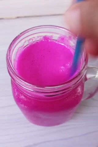 The video shows a close-up overhead view of a clear glass jar filled with a vibrant pink, foamy liquid, likely a smoothie or milkshake. A blue straw is visible, being inserted into the liquid. A hand is partially visible in the upper left corner, out of focus and blurred, suggesting movement as it manipulates the straw or jar. The jar has a handle and is sitting on a light-colored, textured surface that appears to be wooden planks. The lighting is bright, casting highlights on the liquid and the glass. The overall impression is a simple, clean shot focusing on the texture and color of the drink.