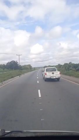 The video shows a vehicle driving on a highway with a white pickup truck initially in front. The pickup truck moves into the opposite lane to pass. The highway is asphalt with white lane markings and yellow edge lines. The surrounding environment consists of green vegetation and trees on both sides of the road. Power lines run along one side of the road. The sky is partially cloudy.