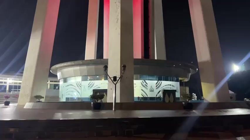 The video shows a low-angle, nighttime shot of a large, modern monument. The monument consists of several tall, angled concrete pillars that support a circular structure at the base, which appears to be a building with large glass windows. Vertical red lights are illuminated on the interior of the monument, creating a dramatic effect against the dark sky. The ground in front of the monument is paved, and there are some potted plants visible. The camera is static for the duration of the clip.