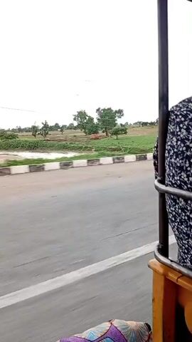 A video clip shows a view from inside a moving vehicle, likely an auto-rickshaw, looking out at a rural landscape. The road is paved and shows a white line marking the lane. On the right side of the road, a concrete barrier with square segments runs along the side. Beyond the barrier, there are fields of green crops and some trees. In the distance, a small hill or mountain is visible under a cloudy sky with visible power lines overhead. The vehicle's interior is partially visible at the bottom of the frame, showing a patterned fabric seat. The scene suggests a journey through a rural area, possibly in India, given the presence of the auto-rickshaw.