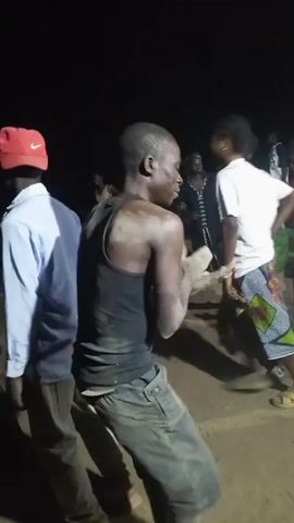 At night, a group of people is gathered in a sandy area, and one man in the foreground, wearing a black tank top and dirty pants, is dancing. He is covered in dirt or dust, and his movements are energetic. Other people surround him, some also dancing. A man in a light blue shirt and red baseball cap stands to the left, while others wear green shirts or white tops. The scene is dark, illuminated only by artificial light sources.