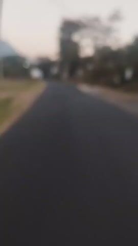 A blurred video shows a dark asphalt road stretching forward. On either side of the road are grassy areas and sparse vegetation. In the distance, trees and buildings are visible, along with mountains under a pale sky. A utility pole with wires runs along the side of the road.