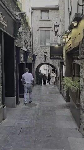The video shows a narrow cobblestone alleyway with stone buildings on either side. Several people are walking in the alley. On the left, a man in a blue shirt and jeans walks away from the camera. To his right, there is a dark storefront with signage. Further down the alley, an archway leads to another section. In front of the archway, a woman in a light-colored dress walks towards the camera, looking at her phone. A group of people are walking through the archway. On the right side of the alley, there are plants and signage for "PETRONELLA" and "BUTTERSLIP LANE". The lighting suggests it is daytime.