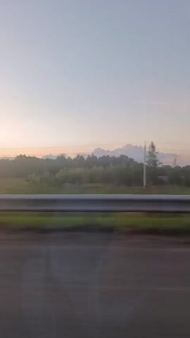 The video captures a view from a moving vehicle, likely a car, looking out the side window. The time of day appears to be either sunrise or sunset, with a soft, pastel-colored sky transitioning from pale blue at the top to hues of orange and pink near the horizon. A line of trees and vegetation forms a dark silhouette against the brightly lit sky in the distance. In the foreground, a guardrail runs along the edge of a road, partially obscuring the view of the landscape. The road surface itself is visible, with a white line marking the lane. The motion of the vehicle creates a slight blur in the foreground, giving a sense of speed and movement. A side mirror of the vehicle is visible in the lower left corner of the frame, reflecting a partial view of the interior and a person's arm. The overall impression is one of a scenic drive through a rural or semi-rural landscape during twilight.