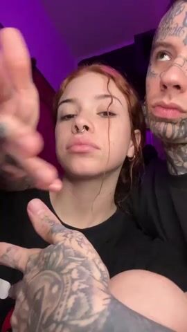 Two people are in the video. A woman with reddish hair and a nose ring, and a heavily tattooed man. Both are wearing black shirts. The man has multiple tattoos on his face, including one resembling a bandage on his cheek. His arms and hands are also tattooed. He is holding up his hands in front of the camera. The background is purple.