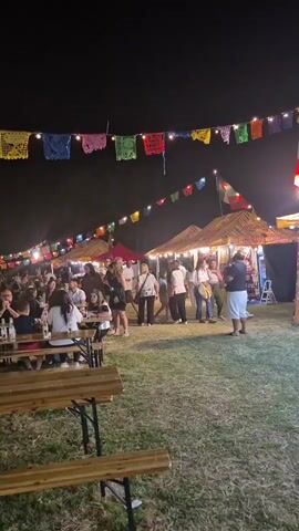 The video shows an outdoor night market or festival. Numerous people are seated at wooden picnic tables in a grassy area. Strings of colorful pennant banners and small lights are strung across the scene. In the background, there are tents or stalls, also illuminated, and more people are visible milling around. The atmosphere appears to be festive and social.