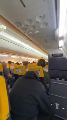The video shows the interior of an airplane cabin filled with passengers seated in rows. The seats are upholstered in a combination of yellow and blue. Overhead, there are lights, ventilation nozzles, and an exit sign. The camera shakes momentarily, then a man wearing a black leather jacket and a hat looks into the camera, touching his face with his finger. The camera returns to show the back of the seats and passengers in front.