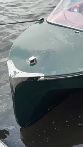 The video is a close-up shot of the front of a dark green boat with a chrome trim at the bow. A small red navigation light is visible on the port side of the bow. Water is present on the deck, creating reflections. The camera pans slightly to reveal the interior of the boat, which features a wooden deck and tan leather seats. The name "La Bulka" is embossed on the back of one of the seats. A person wearing a white shirt and shorts is partially visible in the upper right corner of the frame, standing behind the boat. The water appears calm.