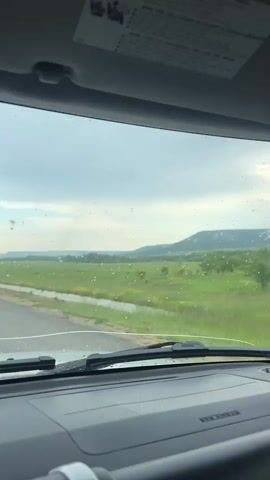 The camera, positioned inside a vehicle, shows a rainy day landscape. The view through the front windshield captures a grassy field adjacent to a road, with a distant tree-covered hill under a cloudy sky. Raindrops dot the windshield. The camera then pans to the right, revealing the car's interior: a white pillar with a handle and an air vent. A side-view mirror shows the road stretching back behind the car, with a yellow line marking the lane. The focus remains on the landscape and the rainy conditions outside the vehicle.