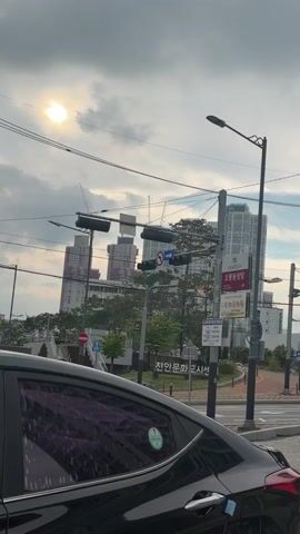 The video shows a street scene with a black car in the foreground. In the background, there are several tall buildings, some of which appear to be under construction with cranes visible. There are traffic lights and street signs, including one with Korean writing. The sky is overcast with the sun peeking through the clouds. There are power lines crisscrossing the frame. Another car is visible in the distance.