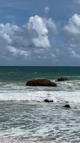 A wide shot of a beach with rough waves crashing against rocks. The sky is blue with large white clouds. The ocean is a deep green and turquoise. The sand in the foreground is a light brown. The waves are white and foamy.