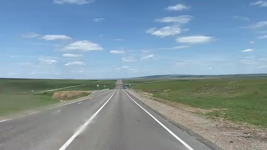 The video shows a view from inside a vehicle driving down a straight, two-lane road. The road stretches into the distance, disappearing over a slight rise. The sky is blue with scattered white clouds. On either side of the road is flat, grassy terrain. There are road signs visible on the sides of the road.  The windshield appears to be slightly dusty.  There are no other vehicles immediately visible in the frame.