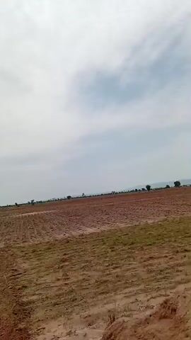 The video captures a panoramic view of an agricultural field under a cloudy sky. The foreground displays freshly tilled red soil with visible furrows, indicating recent plowing. In the mid-ground, a strip of green vegetation, possibly young crops or grass, separates the tilled land from other areas. In the distance, a landscape unfolds with rolling hills, scattered trees, and evidence of human infrastructure such as power lines and possibly fencing along the horizon. The sky is predominantly overcast with a mix of gray and white clouds, suggesting a diffused light condition. The camera appears to be in motion, panning across the expansive rural scenery.