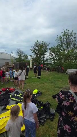 A group of people, including adults and children, are gathered on a grassy field, likely at an event. Two firefighters, one in full gear, are standing in the center, holding and demonstrating something with red balloons. Firefighting equipment is spread out on a sheet in the foreground. The sky is overcast, and trees are visible in the background. There's also a greenhouse-like structure in the distance. People are watching the demonstration.