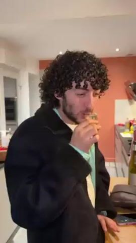 A man with curly dark hair and a beard is standing in a kitchen. He is wearing a black jacket over a light green shirt and a white undershirt. He picks up a small glass, examines it, and puts it down. He is standing in front of a countertop with a brown bag and a bottle of wine on it. In the background, there is a kitchen with white cabinets, a red wall, and other kitchen items. Another person in a red shirt is partially visible in the background. The man appears to be tasting something or cleaning the glass.