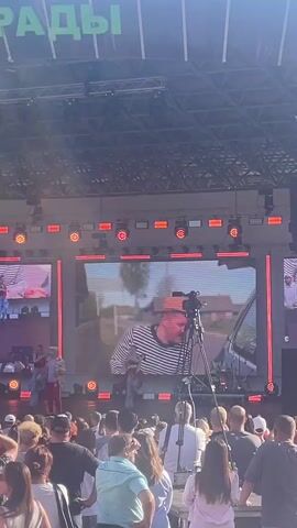 The video shows an outdoor concert or event with a large crowd of people facing a stage. The stage features a large LED screen displaying various video content. On the left side of the screen, a group of performers, including individuals in colorful clothing, are visible on a smaller stage area. On the right side of the screen, a man in a striped shirt and a hat is shown in a close-up shot, appearing to be in a car. A camera on a tripod is positioned in front of the main screen. The crowd consists of people of various ages, many of whom are holding up their phones to record or take pictures. The overall environment appears to be a sunny day, suggested by the lighting and the shadows.
