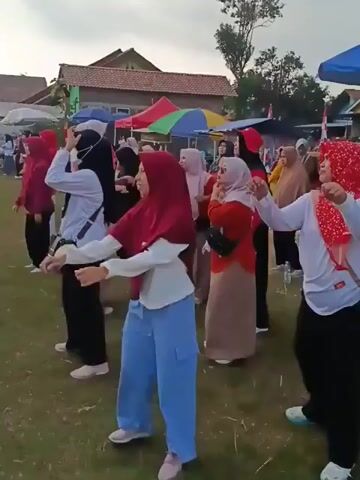 The video shows a group of women exercising outdoors. They are all wearing hijabs and appear to be participating in a synchronized dance or aerobic routine. The setting is a grassy field with several tents and buildings in the background, suggesting an event or community gathering. The lighting suggests it is either early morning or late afternoon.