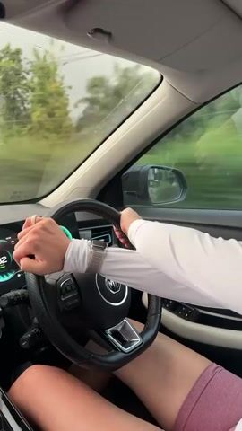 This video shows a person driving a car. The camera is inside the car, looking forward. The driver's hands are on the steering wheel, and their legs are visible. The car's dashboard shows a speed of 84, which then changes to 82. The scenery outside the car is blurred, indicating movement. There are trees and a building visible. The weather appears overcast or rainy, as the windshield has streaks on it.