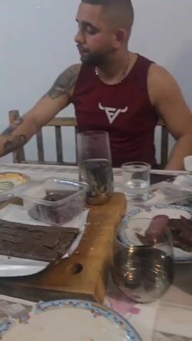 The video pans across a table littered with food and drink remnants, suggesting a recent gathering. Empty bottles and leftover food containers are visible, along with half-eaten plates and serving dishes. One person is at the table, with a tattooed arm resting near the clutter. The setting is a room with a brick wall, a wooden ceiling, and what appears to be a television playing in the background. The atmosphere suggests a relaxed, post-celebratory setting.
