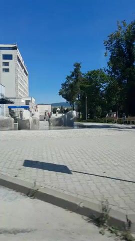 A blue hatchback car is parked in a lot. A white sedan is driving past. A brown SUV is parked in a lot. A person is standing near a parking sign. Another car is parked in a lot. A modern white building with windows and balconies is visible. The sky is blue and clear.