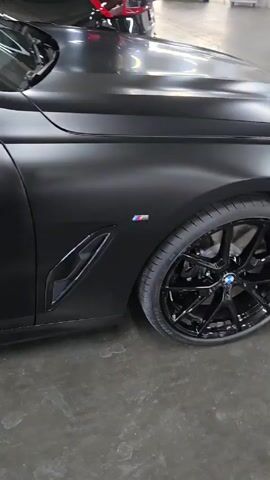 The video shows a matte black car parked indoors. The camera slowly pans along the side of the car, showing the details of the body, door handle, tires and wheels, and taillight. The interior of the car is visible through the window and appears to be red. A person's face is briefly visible in the reflection on the car window.  The floor is gray and appears to be concrete. Other cars are visible in the background.