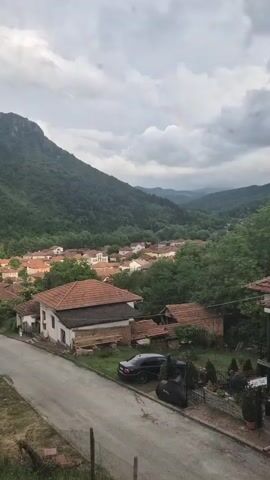 This video captures a panoramic view of a village nestled within a lush, green valley, surrounded by forested mountains under a cloudy sky. The scene is dominated by numerous houses with distinct red-tiled roofs, scattered amidst dense greenery. A paved road is visible in the foreground, with a black car and a covered motorcycle parked on its side.

The camera slowly pans right, revealing more of the sprawling village and the expansive valley beyond. The mountains rise steeply on either side, covered in a rich canopy of trees. In the distance, a faint rainbow arches across the overcast sky, suggesting recent rain or lingering moisture in the air. The overall impression is one of a peaceful, rural setting in a mountainous region, characteristic of Lesidren, Bulgaria. No people or text are visible in the frame.