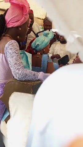 Here's a concise description of the video:

The video shows a scene at what appears to be a wedding or celebratory event. A couple is seated in a decorated carriage or seat, surrounded by other people, some of whom are taking pictures with their phones. The women in the video are dressed in brightly colored traditional attire, and many are wearing headwraps.  The setting looks like it might be outdoors under some kind of shelter. There is music or speech audible in the background.