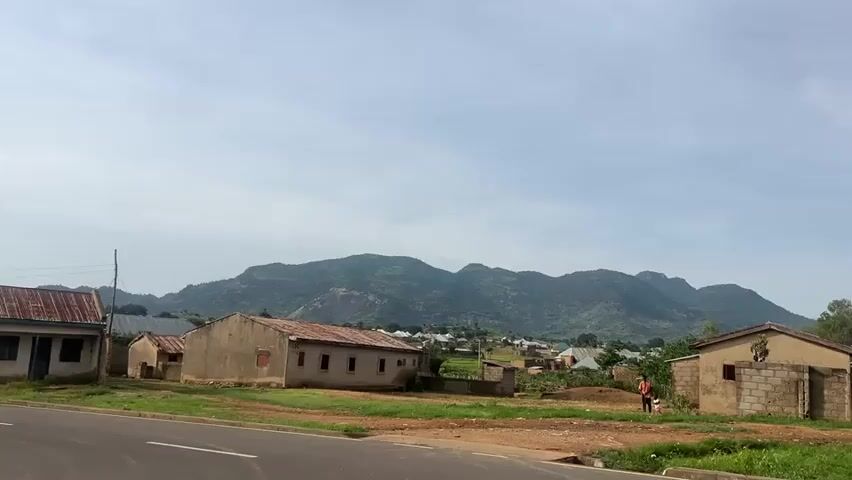 A wide shot shows a rural African village with simple houses and a paved road in the foreground. The houses are mostly made of mud brick with corrugated iron roofs, some appearing unfinished. A person is walking on the side of the road near the houses. In the background, a range of green, hilly mountains stretches across the horizon under a partly cloudy sky. The overall impression is one of a dry, rural landscape.
