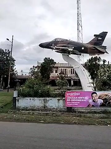 The video shows a static display of a military jet on a pedestal. The jet is painted in camouflage colors. Behind the jet is a multi-story building with a flat roof and many windows. A communication tower is visible in the distance, behind the jet.  A white concrete structure supports the jet. There is a low wall around the base of the display, with some plants growing inside. A street runs in front of the display. There's a streetlight on the left side of the frame. The sky is overcast. A vehicle is parked near the building. The ground appears to be wet.