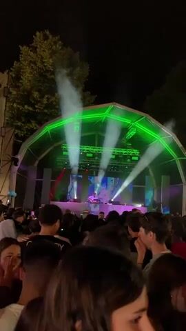 The video captures a lively outdoor concert at night, featuring a DJ performing on a large stage. The stage is illuminated with bright green lights and a massive LED screen displaying dynamic blue and white visuals, including images of rocks and a horned figure resembling a Viking. The DJ, dressed in a white shirt, is actively mixing music at a console on the stage. Below the stage, a crowd of people is visible, with some individuals turning their heads towards the camera. The atmosphere is energetic, with smoke effects rising from the stage and beams of light sweeping across the scene.