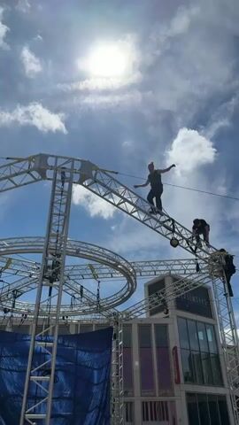 The video shows three people standing on a high metal structure with a sky backdrop. A fair-skinned person with a ponytail raises their arms in the air while standing on a slanted section of the structure. Two other people are nearby; one is on their hands and knees, and the other is sitting with their legs dangling over the edge of the structure. The structure is made of metal beams and supports, and has some circular pieces. A blue tarp is visible underneath part of the structure, and a building with a sign that reads “Glassworks” is visible in the background. The sky is blue with a few white clouds and a bright sun.