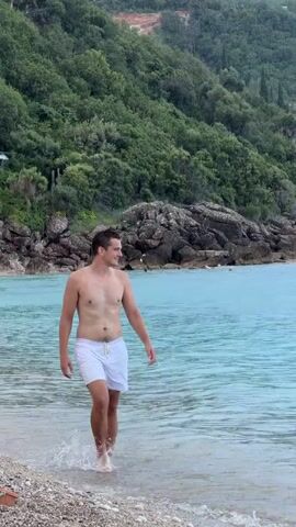 This media file depicts a seaside scene in Drobnići, Montenegro. The primary subjects are two young males and one adult male. The adult male, positioned centrally, is walking out of the turquoise water and onto a pebble beach, wearing white swim trunks. He has a relaxed expression and appears to be enjoying the moment.

To the left and foreground, a younger male is partially visible, looking down and holding snorkeling gear. His body language suggests he is about to engage in an activity. In the background, behind the adult male, a lush green, tree-covered hillside rises steeply, dotted with rocky outcrops. The sea is calm and clear, reflecting the overcast sky. The lighting indicates it is likely late afternoon or early evening, with diffused light suggesting cloudy weather.

A small, light-colored dog with brown markings is also present on the beach in the foreground, facing towards the water. The overall atmosphere is casual and recreational, hinting at a day spent by the sea. There is no visible text within the media file.
