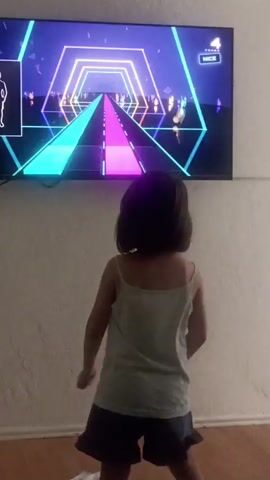 The video shows a young girl with dark hair, wearing a light-colored tank top and a dark skirt, playing a rhythm video game. The game is displayed on a large flat-screen television mounted on a textured, light-colored wall. The game's interface features a neon, geometric tunnel with a path of illuminated footsteps. The girl is facing the television and appears to be moving in time with the game's prompts, with her arms in motion. On the screen, a silhouette of a figure is visible in a small box on the left, likely representing the player's avatar. Text on the screen indicates "COMBO 4" and "NICE." The scene takes place indoors, likely in a living room or play area, with a wooden floor visible at the bottom of the frame. The lighting is typical of an indoor setting, with the game's bright neon lights being the dominant illumination. The overall mood seems to be one of playful engagement with the video game. There are no visible indications of time of day, weather, or specific location within Bağcılar, Turkey.