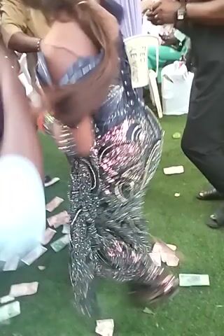 The video shows people in what appears to be a party setting. A woman in a sparkly outfit is seated and dancing as money is thrown around her. Several individuals are visible around her, some dressed in casual attire and others in more formal wear. The ground is covered with what seems to be artificial grass and scattered currency. The atmosphere looks festive and celebratory.