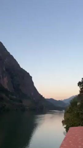 The video shows a scenic view of a body of water, likely a river or lake, surrounded by mountains. The mountains are covered in lush green vegetation and appear to be rocky in some areas. The sky is a clear, pale blue, with a hint of orange and pink near the horizon, indicating it might be dawn or dusk. The water is calm and reflects the colors of the sky and the surrounding landscape. A bridge is visible in the distance, spanning across the water. The video is taken from a slightly elevated position, potentially a pier or a similar structure. There are no people visible in the video.